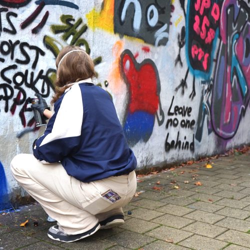 Eine Person in Rückenansicht hockt mit einer Spraydose in der Hand vor einer Wand voll bunter Graffiti und sprüht schwarze Buchstaben daran.