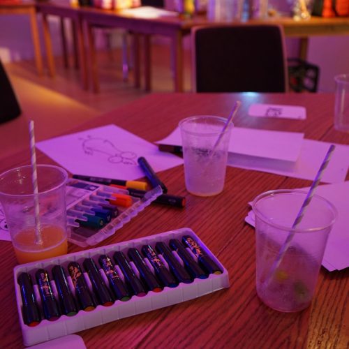 Bild: Auf einem Tisch liegen Stifte und Blätter, einige davon sind bemalt, dazwischen stehen halb leer getrunkene Plastikbecher mit Strohhalmen darin.